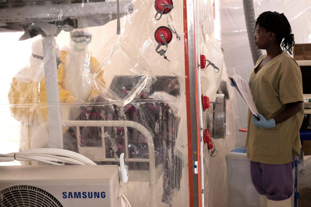 A female healthworker watches as two healthworkers in Ebola protective suits treat a patient.